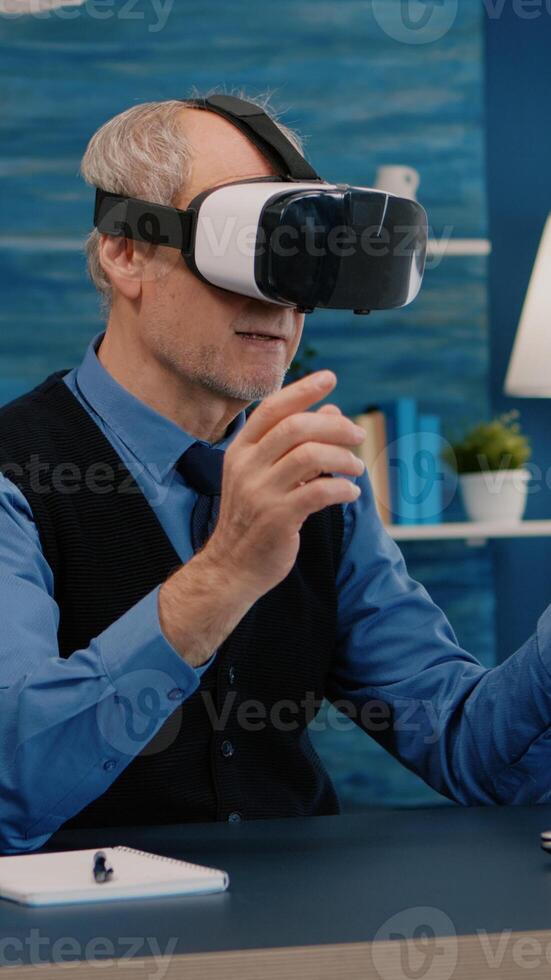 Retired man experiencing virtual reality using vr headset in living room working from home. Old remote employee searching, analysing financial reports while elderly wife watching tv in background photo
