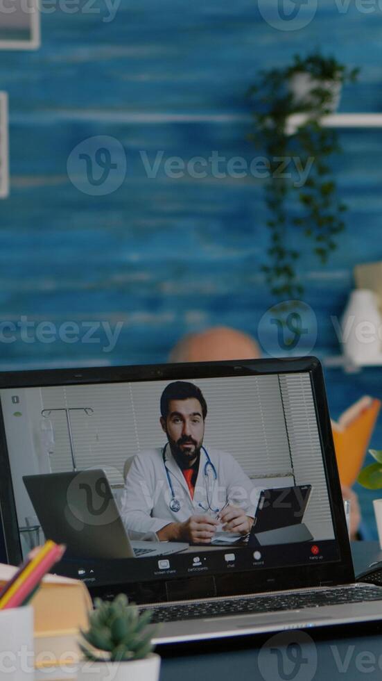 Old woman sitting at table at home looking at laptop answering question about health talks with therapist by videoconference call application, holding pills flask Remote medical consultation concept photo
