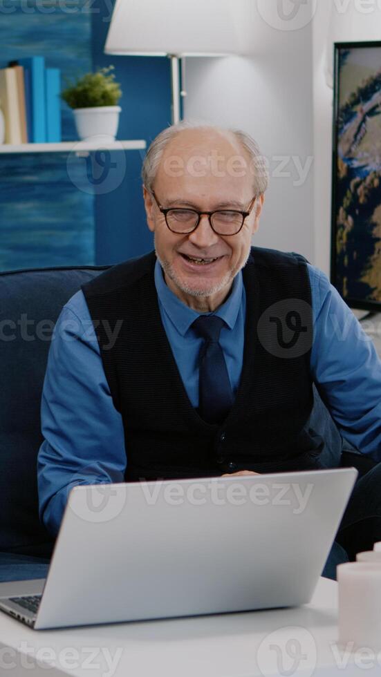 Enthusiastic couple of pensioners sitting together at home on sofa using laptop talking on call with family friends waving at camera during online meeting. Connection lifestyle concept in quarantine photo