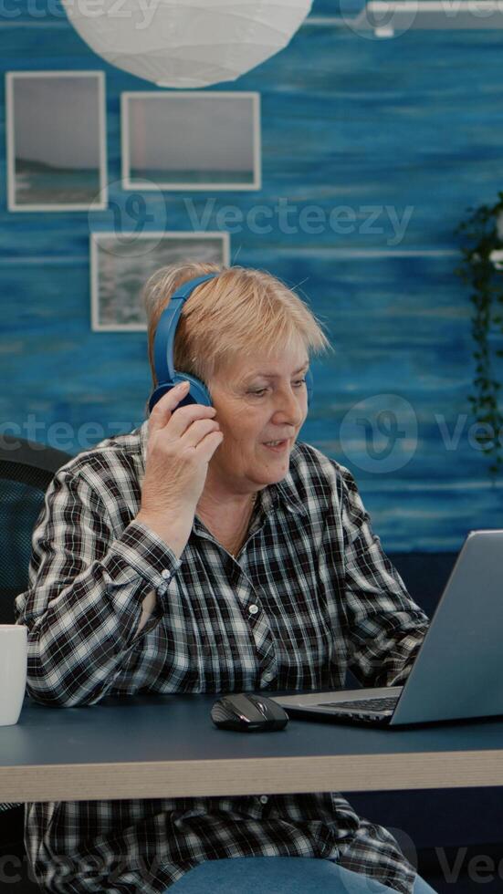 Happy old woman entrepreneur with headset listening music while checking statistics, writing on laptop working from home. Retired worker using modern technology reading typing, searching on computer photo