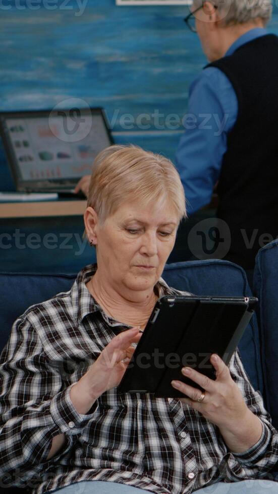 Middle aged old woman relaxing holding tablet reading e book sitting on sofa at home, while senior adult man working at laptop in background. Person enjoying using notepad browsing internet shopping photo
