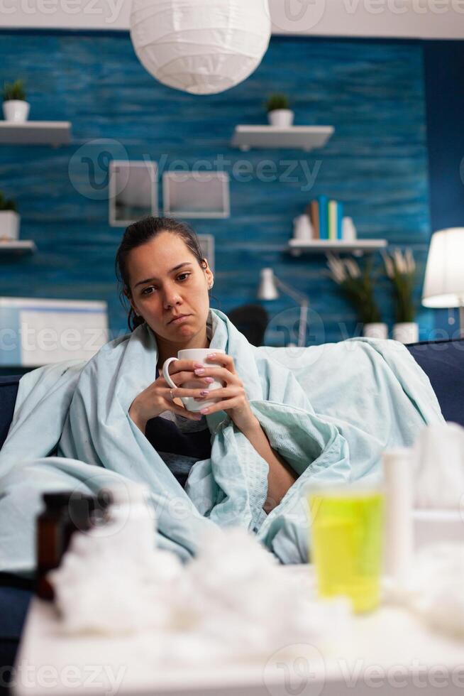 Sick caucasian woman sitting in bed with severe cold and flu drinking hot tea. Adult wrapped in blanket on the sofa with health fever infection symptomps. Person with disease at home, temperature and cough photo