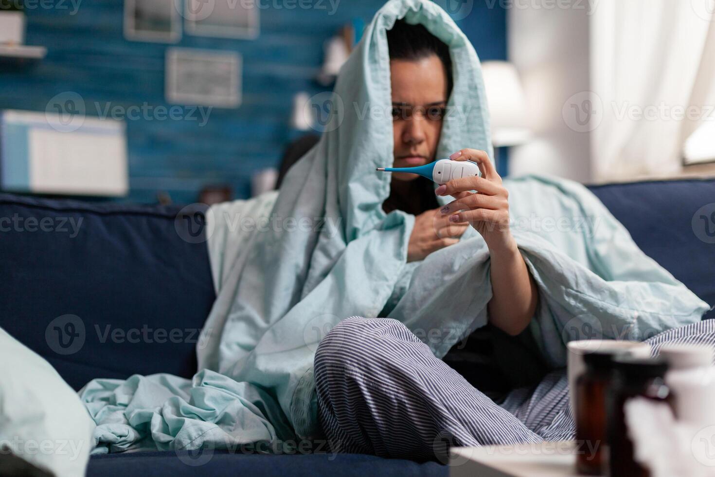 Caucasian woman measuring temperature ill at home with a thermometer. Person feeling sick cold unwell, checking fever and symptoms of disease flu infection. Resting adult with headache photo
