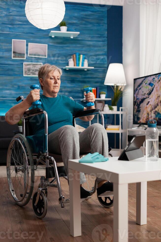 Handicapped senior woman holding trainer dumbbells doing arms exercise working at body persistence training in living room. Disabled pensioner looking at online muscles workout course on tablet photo