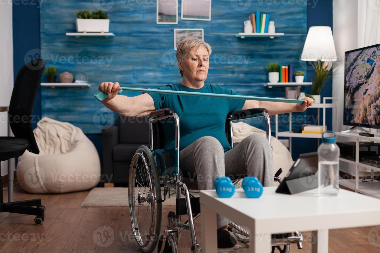 Invalid old woman in wheelchair stretching arm resistance training body muscle using elastic band after leg accident watching therapy on tablet. Pensioner exercising gym workout in living room photo