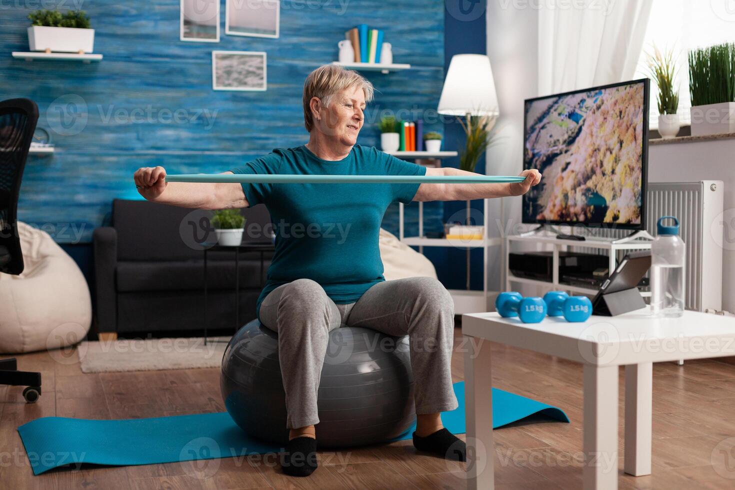 Retired senior man sitting on fitness swiss ball in living room doing wellness fitness workout streching arm muscles using aerobics elastic band. Pensioner training body resistance in living room photo