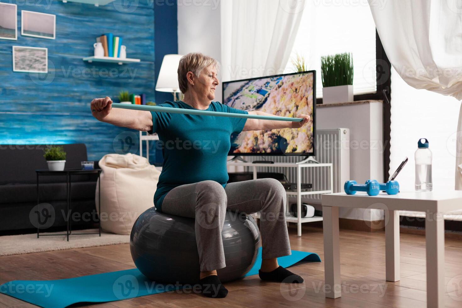 alegre retirado pensionista trabajando brazo músculos utilizando elástico banda practicando aerobio ejercicio. pensionista sentado en suizo pelota en vivo habitación trabajando a cuerpo cuidado de la salud resistencia acecho gimnasio foto