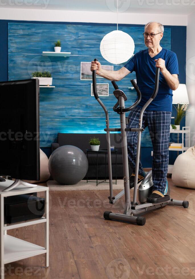 Focused retirement senior man working at legs resistance using cycling bicycle machine watching aerobics on television for well being. Pensioner training body muscle during health cardio workout photo