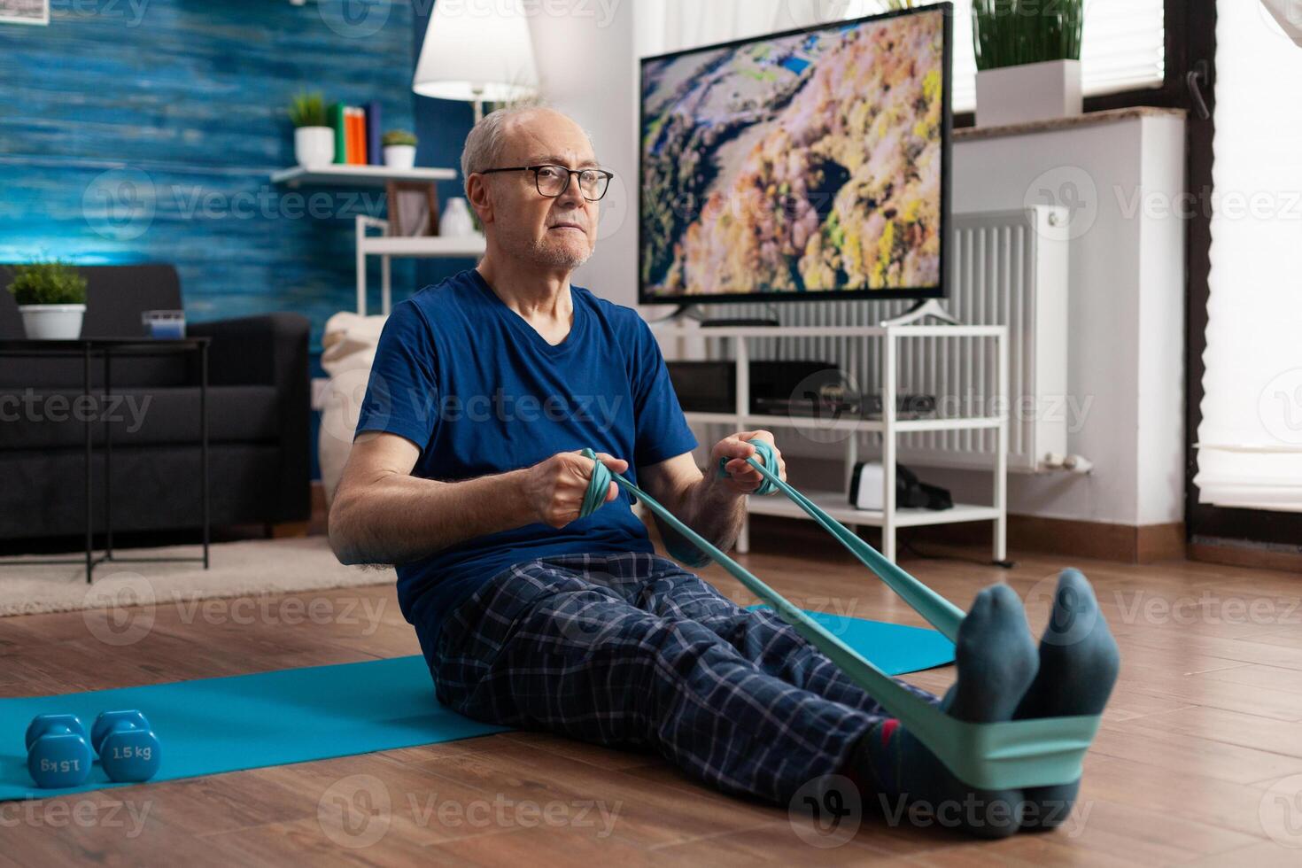 Jubilación mayor hombre sentado en yoga estera extensión piernas músculos utilizando resistencia elástico banda formación cuerpo flexibilidad. pensionista en ropa de deporte adelgazar peso durante músculo formación en vivo habitación foto