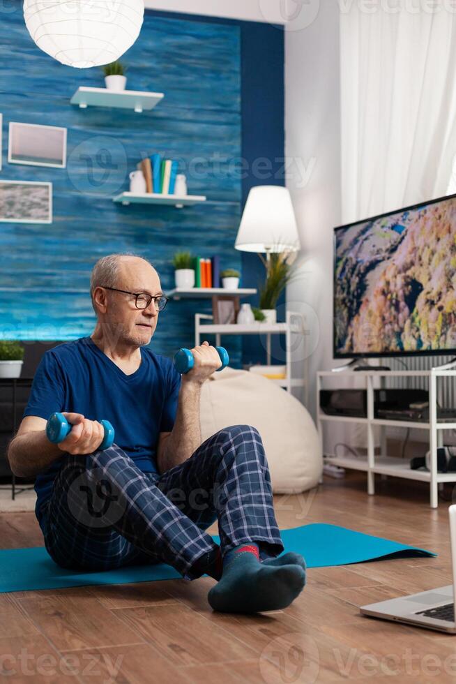 Senior man in sportwear warming up body muscles practicing wellness exercise using workout dumbbells. Pensioner sitting on yoga mat with crossed legs during aerobics routine looking at gym photo