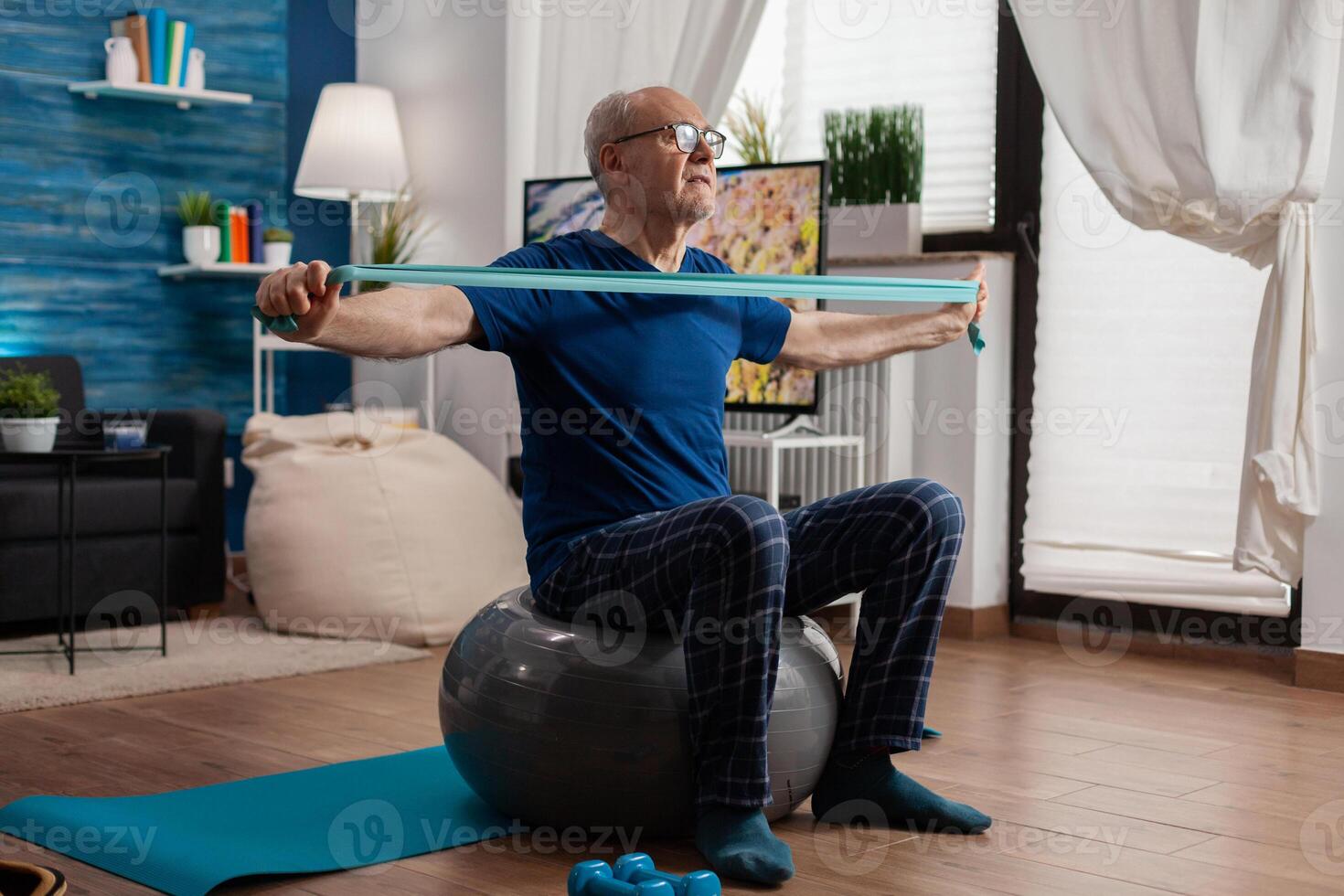 Retiree senior man sitting on swiss ball in living room doing healthcare fitness exercises streching arm muscles using resistance elastic band. Pensioner training body strength in living room photo
