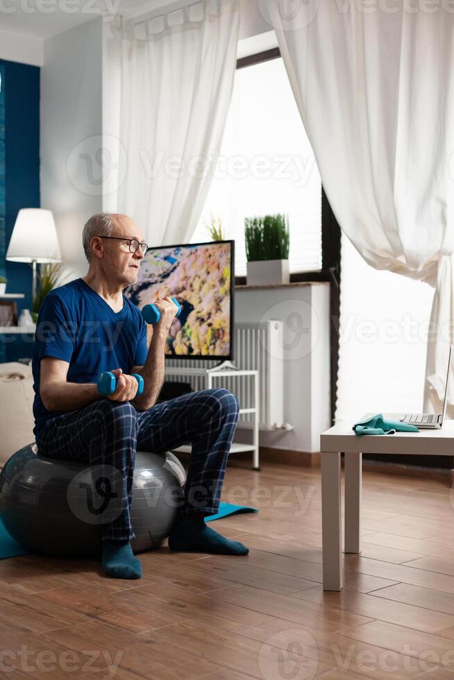 Senior man training body resistance doing arms exercise sitting on swiss ball in living room. Pensioner watching online wellness training on laptop exercising muscle workout using dumbbell photo