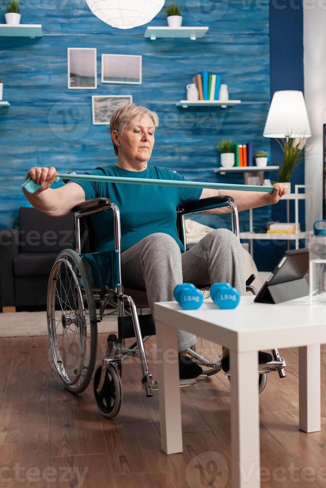 Invalid senior woman in wheelchair holding resistance elastic band stretching body muscle recovering after disability accident watching workout on tablet. Pensioner doing aerobics exercise photo