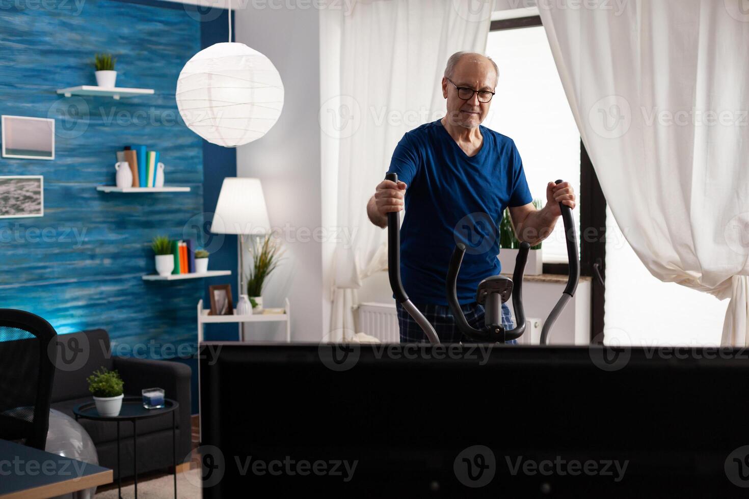 Pensioner doing legs exercise training body muscle using cycling bicycle machine during aerobics workout. Retirement active senior man watching fitness on television in living room photo