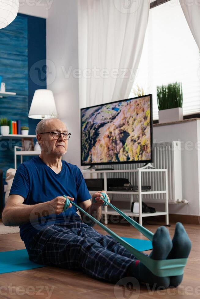 Jubilación mayor hombre sentado en yoga estera extensión piernas músculos utilizando resistencia elástico banda formación cuerpo flexibilidad. pensionista en ropa de deporte adelgazar peso durante músculo formación en vivo habitación foto