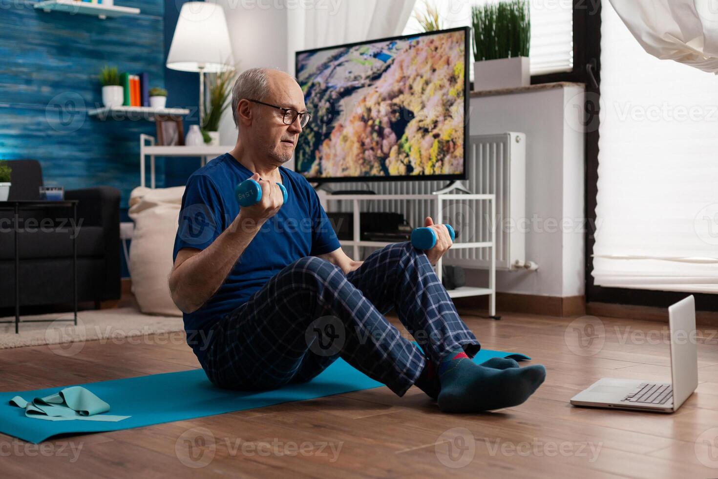 Retired senior man sitting on yoga mat stretching abdominals muscles practicing fitness workout using dumbbells. Pensioner working at body warming up during wellness routine watching pilates photo
