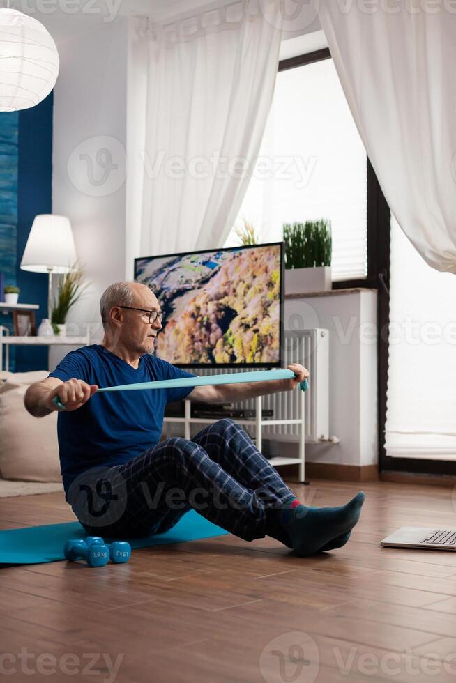 Retirement senior man sitting on yoga mat with leg in crossed position stretching arms muscles using stretch elastic band during sport routine in living room. Pensioner exercising bodyresistance photo