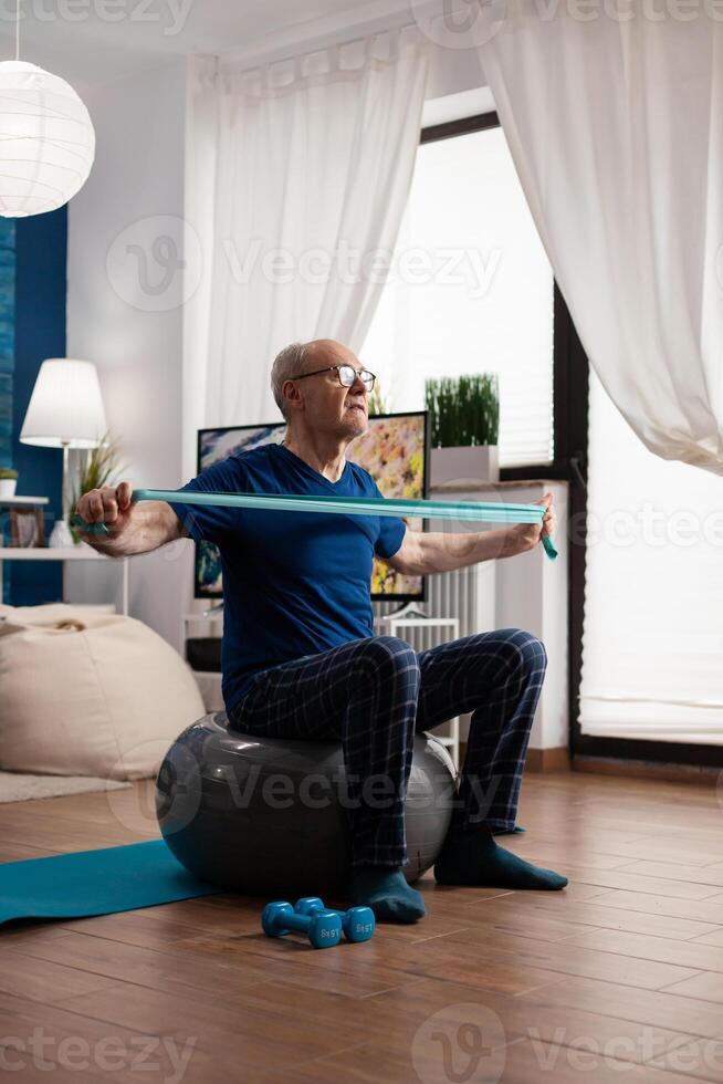 Cheerful retired senior man exercising arm muscles using resistance elastic band practicing aerobic exercise. Pensioner sitting on swiss ball in living room working at body healthcare photo