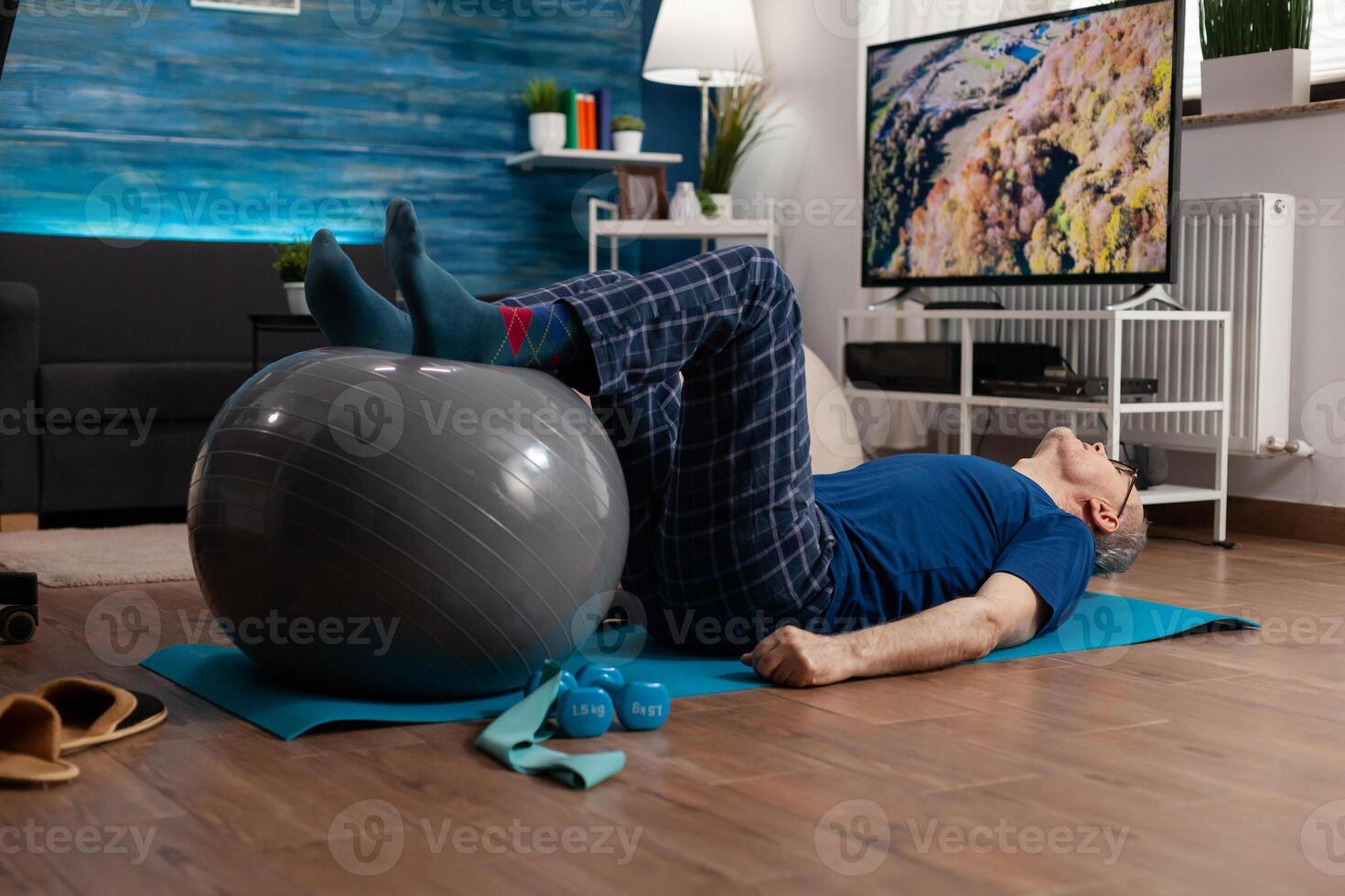 Retirement senior man doing wellness warming legs up using swiss ball sitting on yoga mat in living room. Retired pensioner practicing abdominals exercises working at body muscular healthcare photo
