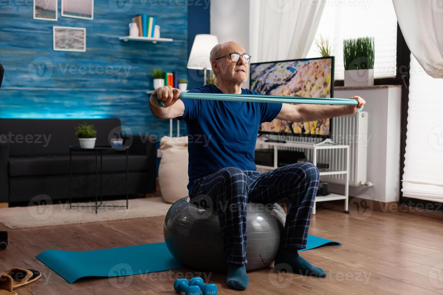 Cheerful retired senior man exercising arm muscles using resistance elastic band practicing aerobic exercise. Pensioner sitting on swiss ball in living room working at body healthcare photo
