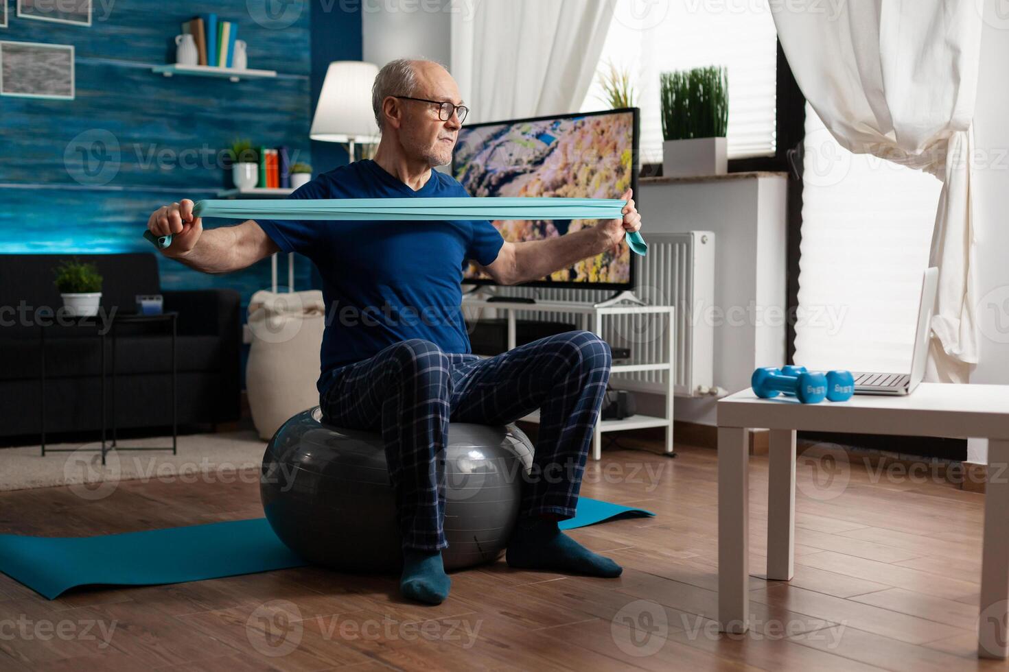 Invalid pensioner practicing active arm exercises using resistance elastic band watching online fitness workout on laptop. Pensioner working body healthcare sitting on swiss ball in living room photo