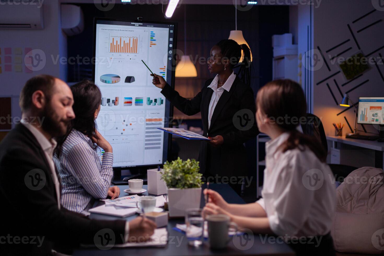 Workaholic african american leader standing in front of presentation monitor explaining marketing project solution late at night in company meeting room. Diverse teamwork working at business ideas. photo