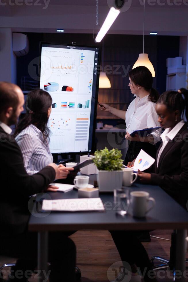 Diverse multiethnic business teamwork overworking in office meeting room analyzing financial graphs late at night. Coworkers brainstorming project ideas solving company problem. photo