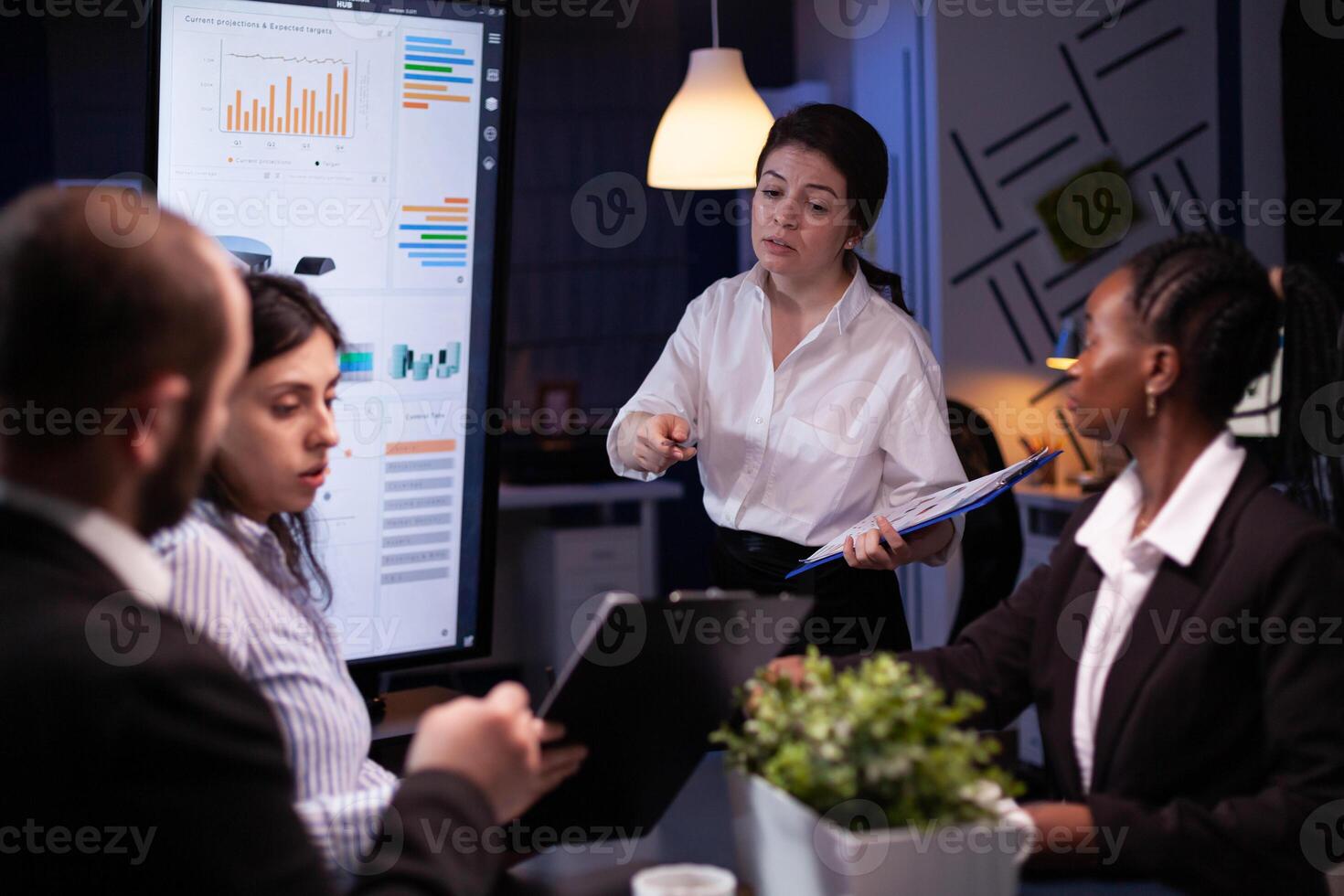 Diverse multi ethnic teamwork brainstorming company ideas analyzing financial graphs presentation. Businesspeople overworking in meeting room late at night discussing solution photo