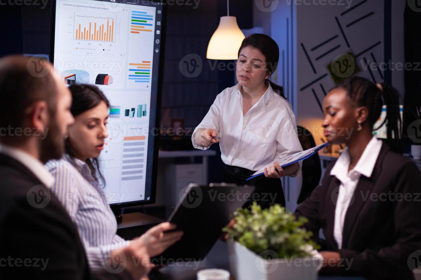 Diverse multi ethnic teamwork brainstorming company ideas analyzing financial graphs presentation. Businesspeople overworking in meeting room late at night discussing solution photo
