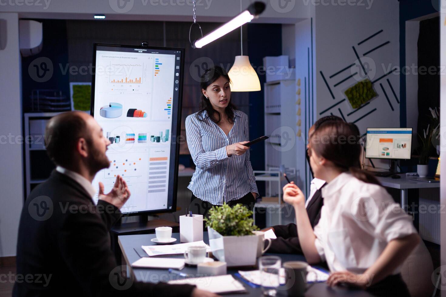 Workaholic businesswoman pointing financial strategy using monitor working overtime in company meeting office room. Diverse multi-ethnic teamwork overworked solving management statistics in evening photo