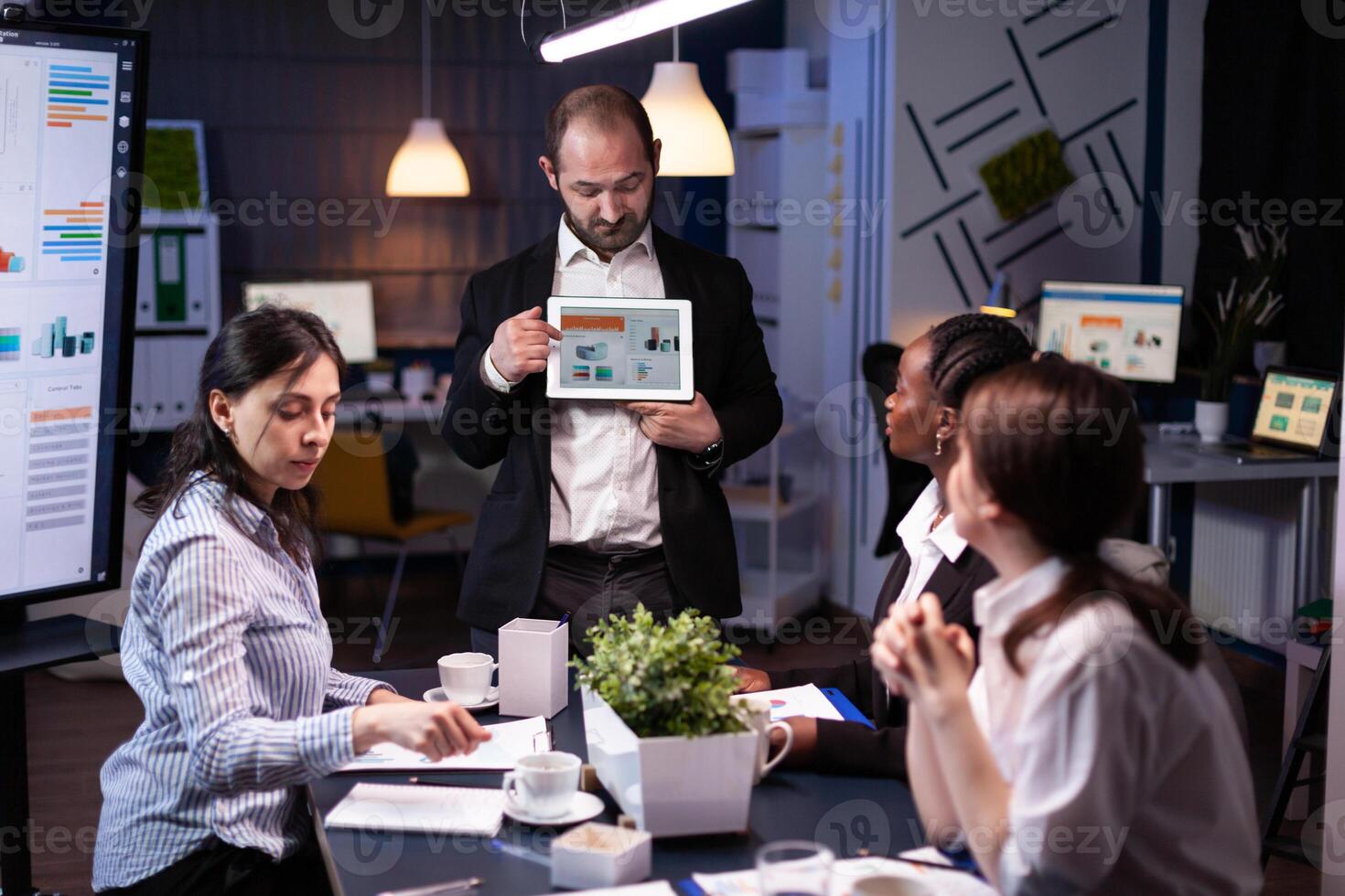 Focused businessman showing corporate graphs presentation using tablet working at company ideas. Diverse multi-ethnic businesspeople brainstorming strategy late at night in office meeting room photo