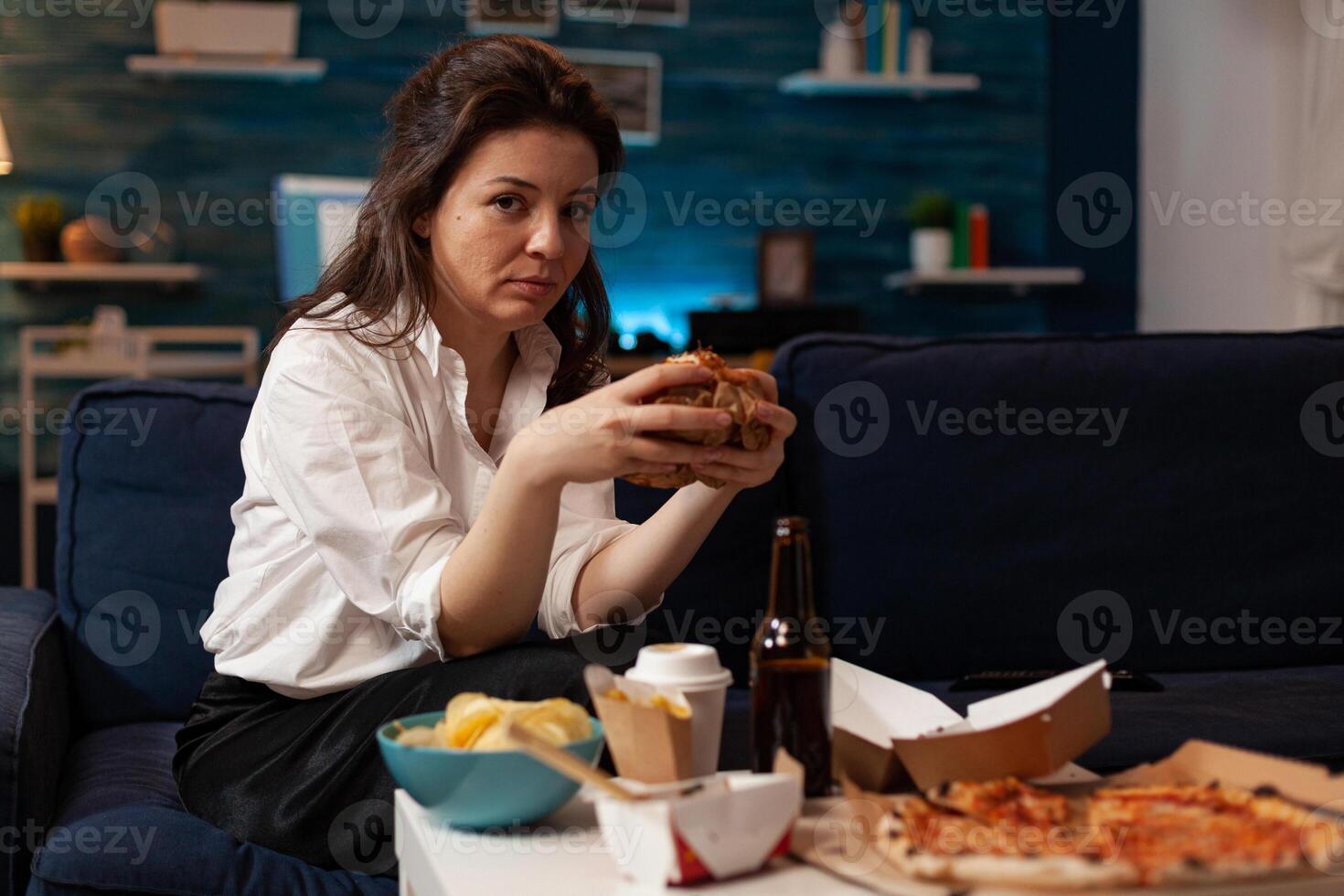Portrait of relaxed woman looking into camera holding tasty burger in hands during fast food night in living room. Caucasian female enjoying delicious home delivered junk-food. Takeaway delivery photo
