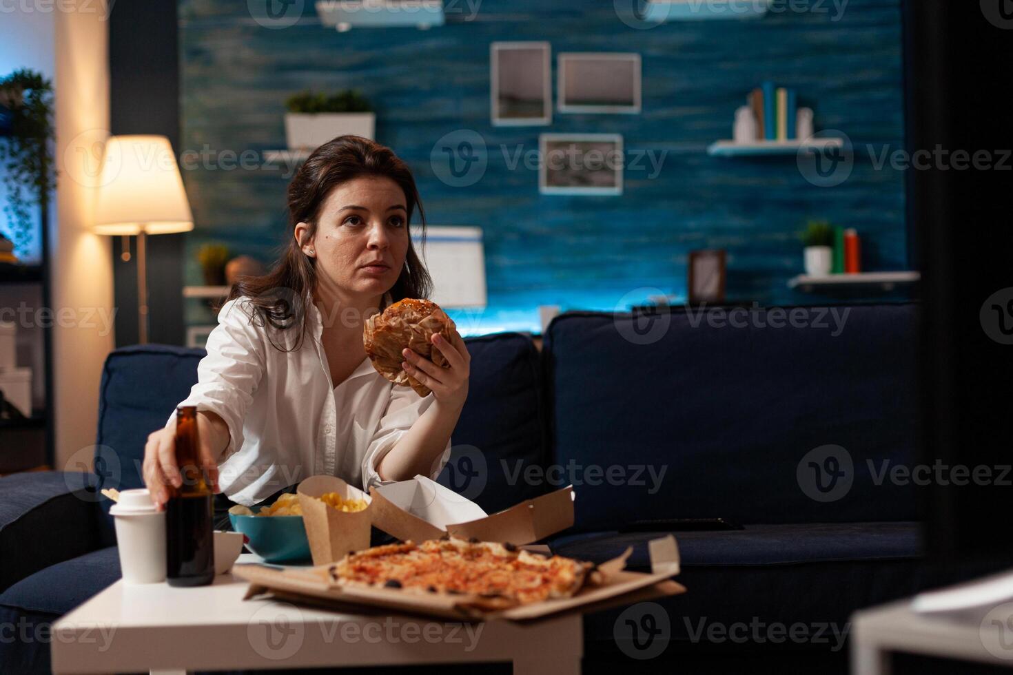 Portrait of woman holding delicious buger eating takeaway delivery food watching entertainment movie on television. Caucasian female sitting on couch tasting tasty junk-food enjoying night photo