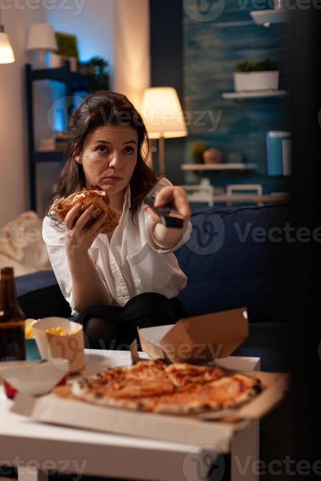 Happy woman changing channels using remote watching entertainment movie on television eating tasty delicious burger in evening. Caucasian female enjoying takeaway food home delivered photo
