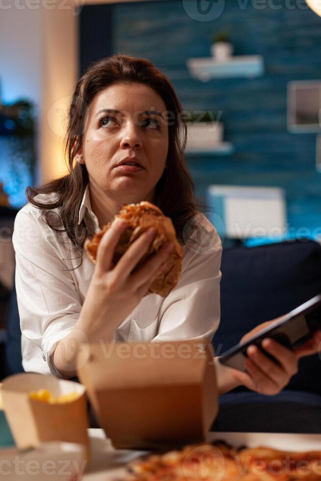 Caucasian female holding tastu burgery in hands browsing on social media using smartphone relaxing on couch during junck-food night. Woman enjoying takeaway food home delivery photo