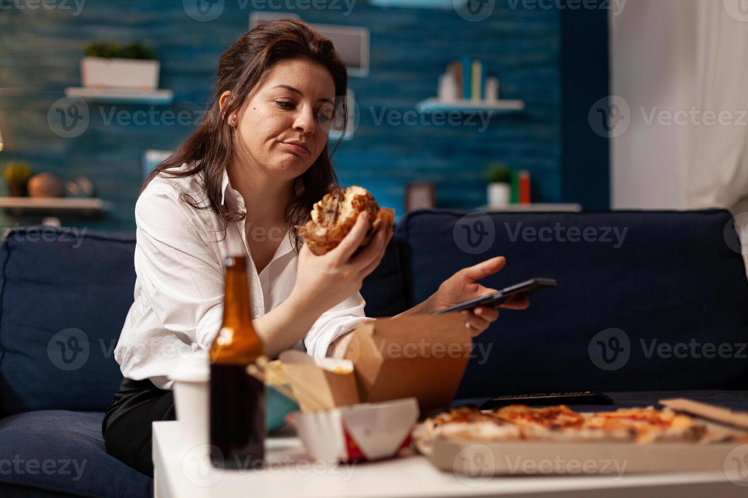 Caucasian female holding tastu burgery in hands browsing on social media using smartphone relaxing on couch during junck-food night. Woman enjoying takeaway food home delivery photo