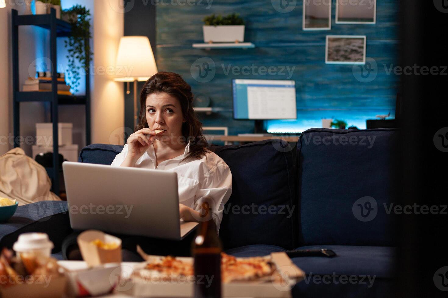 Woman eating tasty snack working on laptop computer while watching documentary movie series on television. Caucasian female enjoying takeaway food home delivered. Fastfood delicious order photo