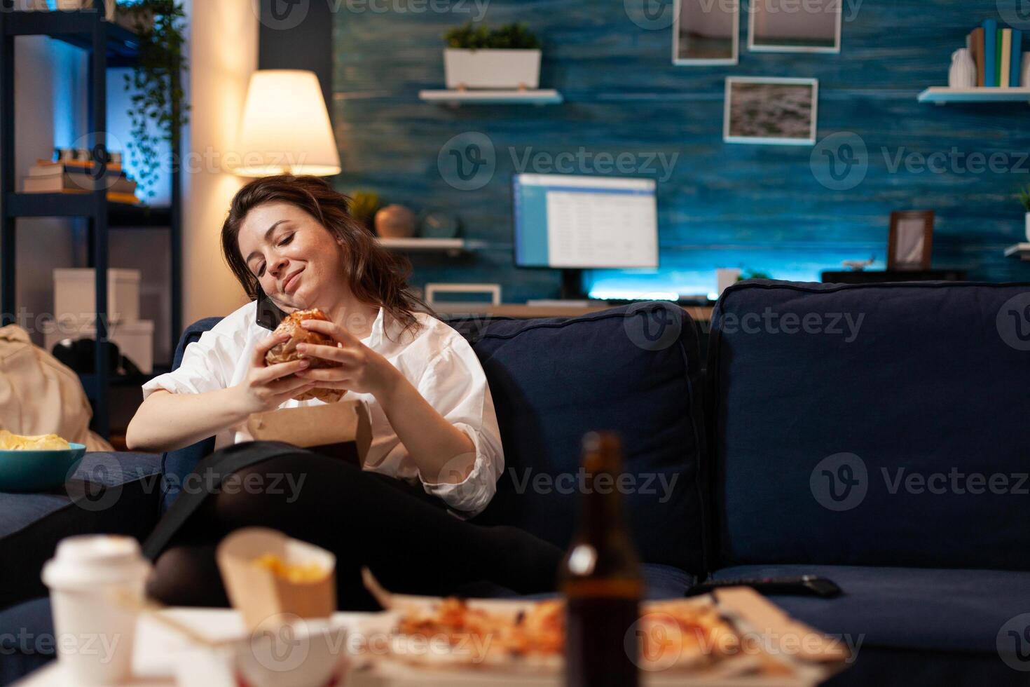 Woman talking at phone with friends while holding tasty delicious burger in hands relaxing on couch in living room late at night. Caucasian female enjoying tajeaway junk food home delivered photo