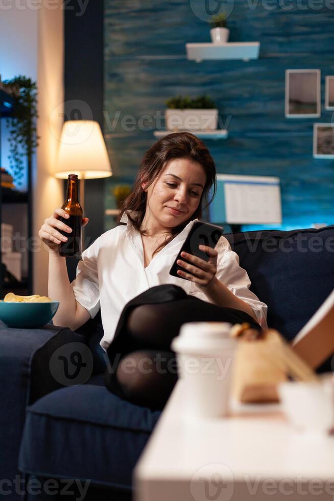 Caucasian female relaxing on couch browsing on social media using smarhphone while drinking beer during relaxing night at home. Woman enjoying takeaway food home delivered. Junk-food order photo