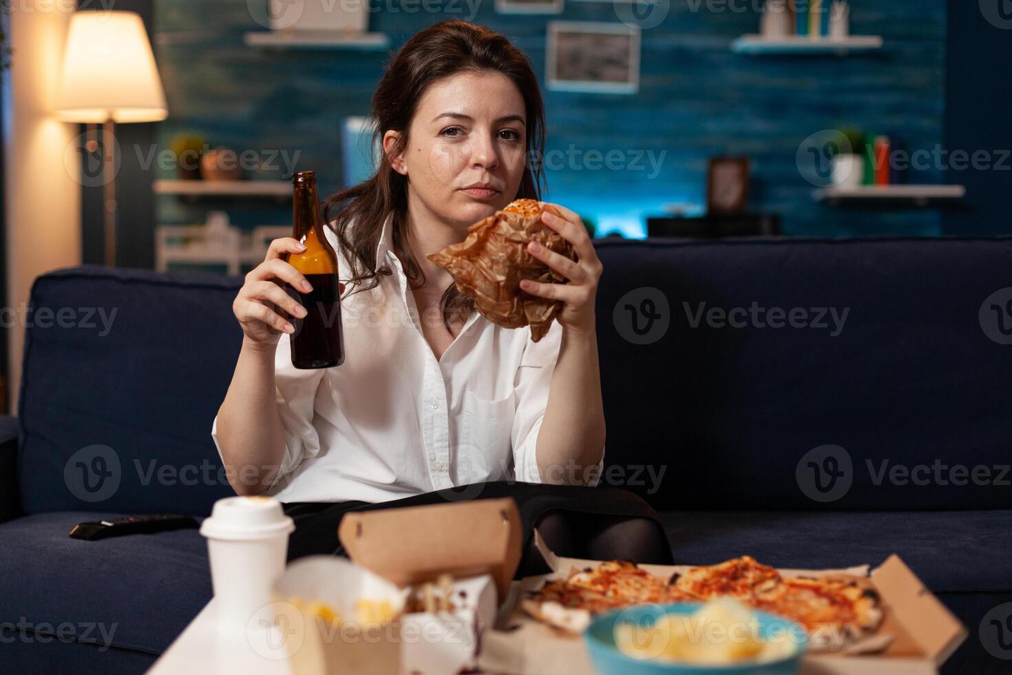 Portrait of caucasian woman looking into camera relaxing on sofa in living room enjoying takeaway food home delivery. Woman eating tasty delicious burger. Fastfood lunch meal order photo