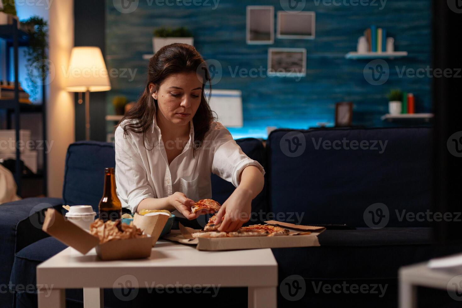 Office worker at home having dinner taking a hot pizza slice from cardboard box on table with delicious hamburger and fries. Woman having takeaway fast food menu and beer in front of television. photo