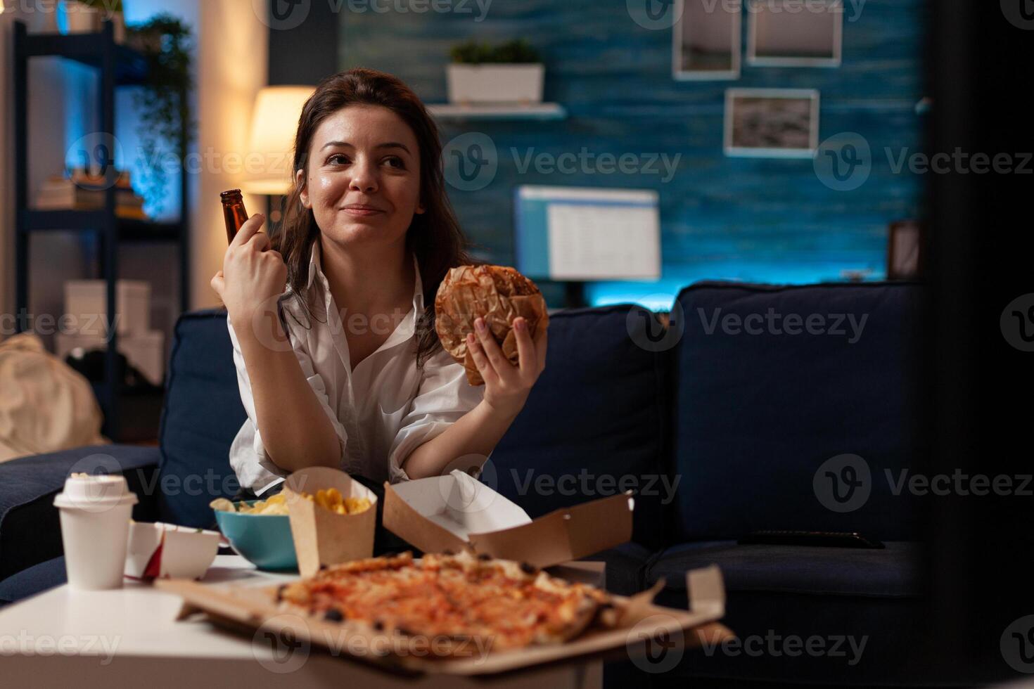 Woman eating tasty takeaway fast food hamburger while enjoying a cold beer in front of television with comedy show in home living room. Smiling person having hot pizza and takeout delivery menu. photo