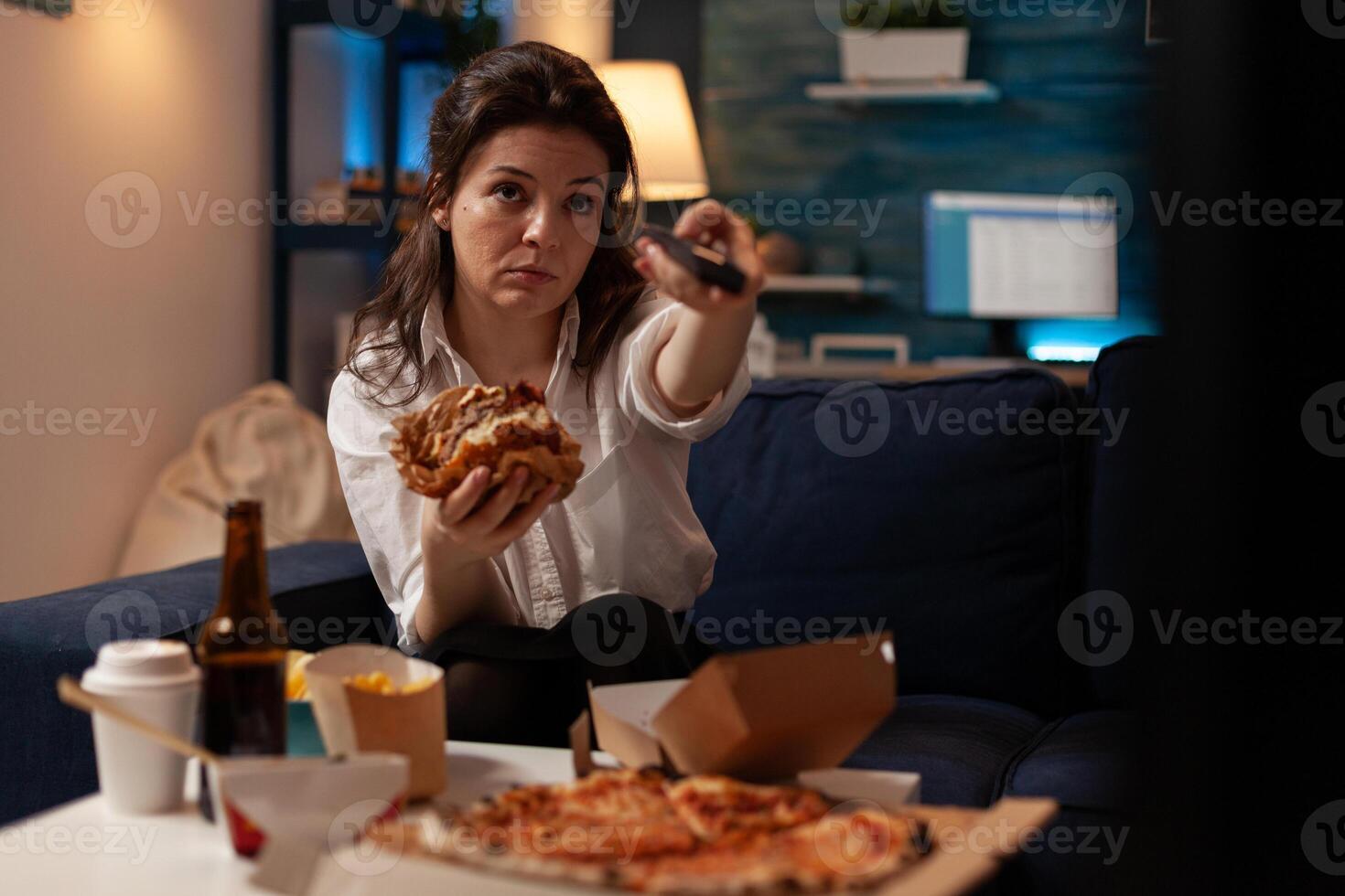 Young woman sitting on sofa eating double cheeseburger holding remote turning up volume on television watching evening news. Person changing tv channels while eating takeaway hamburger. photo