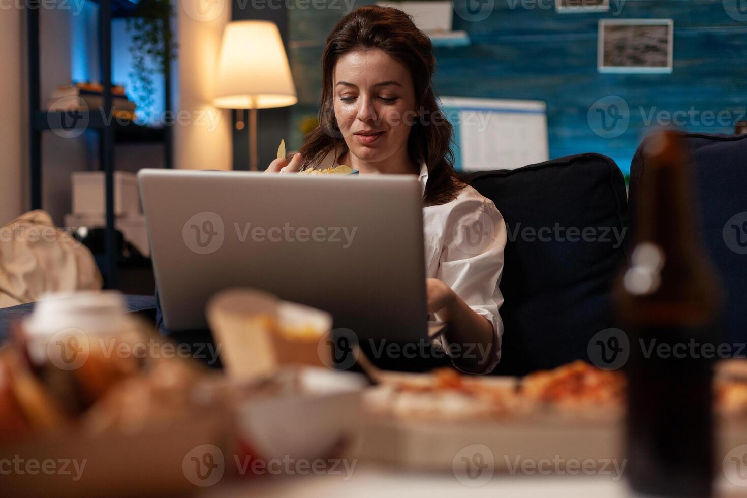 Woman browsing social media on laptop while having fast food snack sitting on couch in living room. Office worker with personal computer eating chips in front of table with home delivery menu. photo