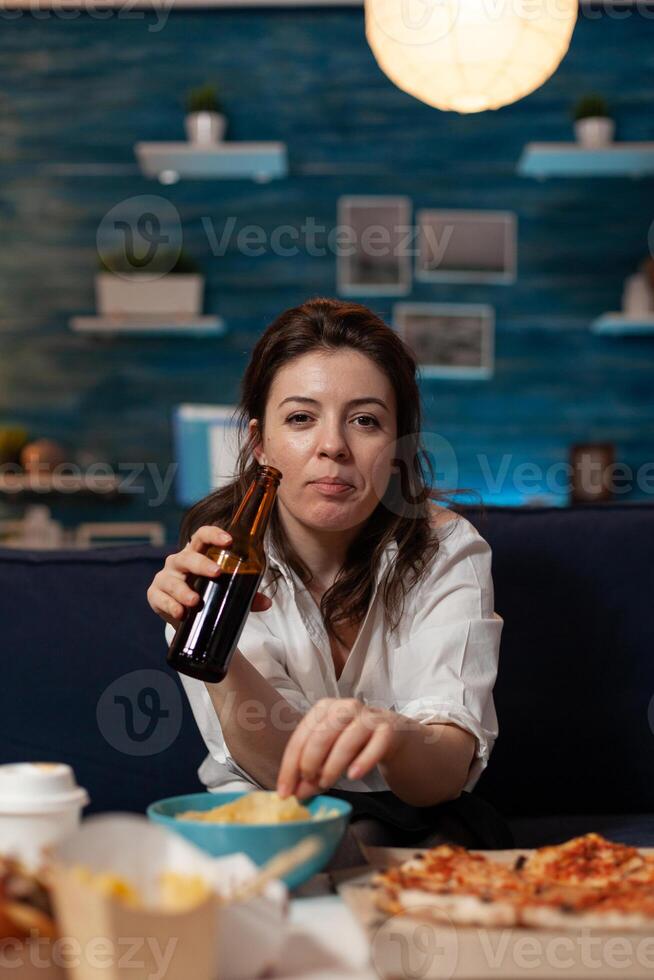 Portrait of woman looking at camera holding beer bottle and eating potato chips from bowl at table with large pizza and takeaway dinner. Office worker on couch relaxing after takeout menu. photo
