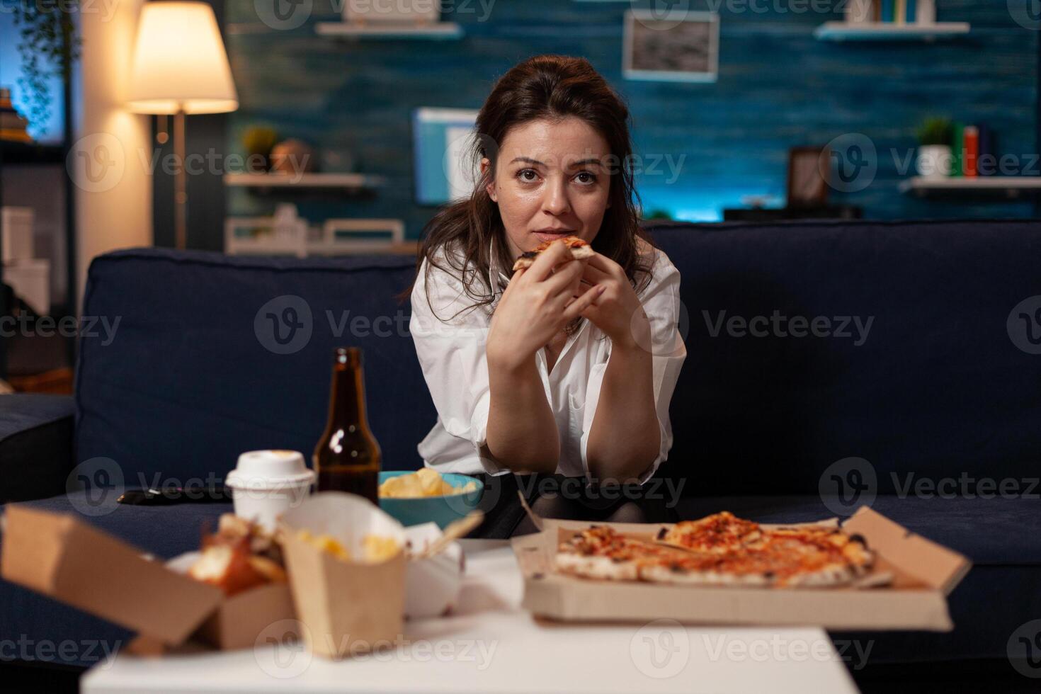 Person sitting on couch in living room having a slice of delivery pizza while watching television sitcom at table with fast food takeout menu. Woman after office work eating takeaway tv dinner. photo