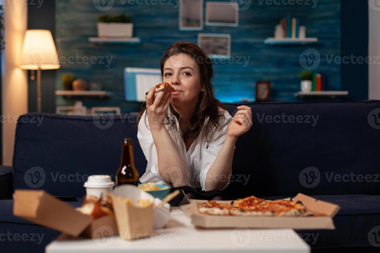 Woman after office work taking a bite out of slice of hot delivery pizza while watching television at table with fast food takeway menu. Person sitting on couch in living room eating takeout dinner. photo