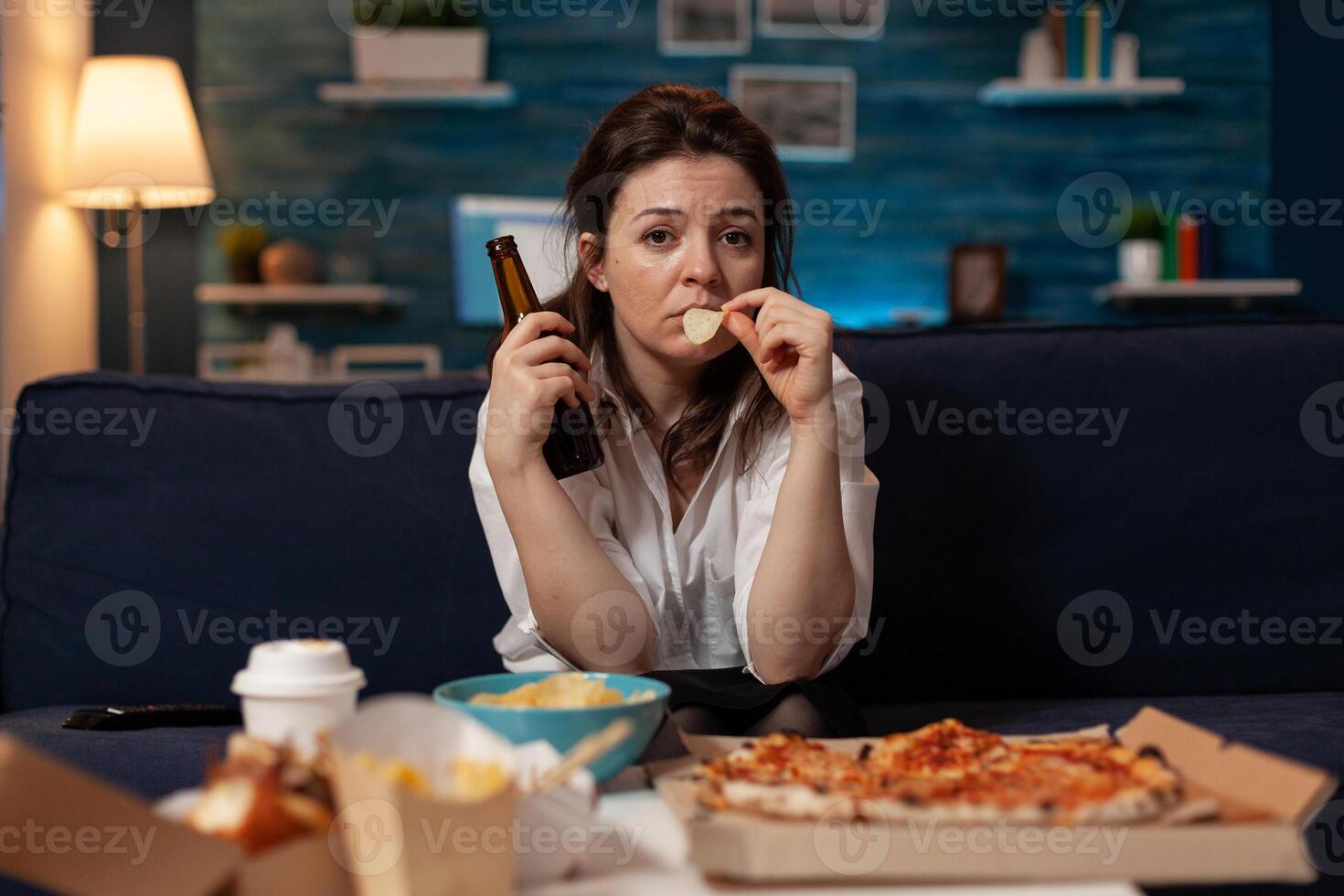 Sad woman drinking craft beer and eating potato chips from bowl at table with large pizza and takeout dinner in home living room. Tired office worker on couch relaxing after takeaway menu. photo