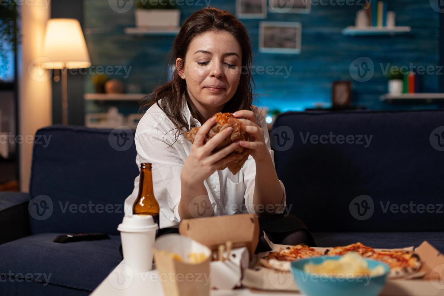 Woman sitting on couch looking at tasty delicious burger delivery dinner in living room in front of television. Person standing at table with takeaway fast food meal large pizza. photo