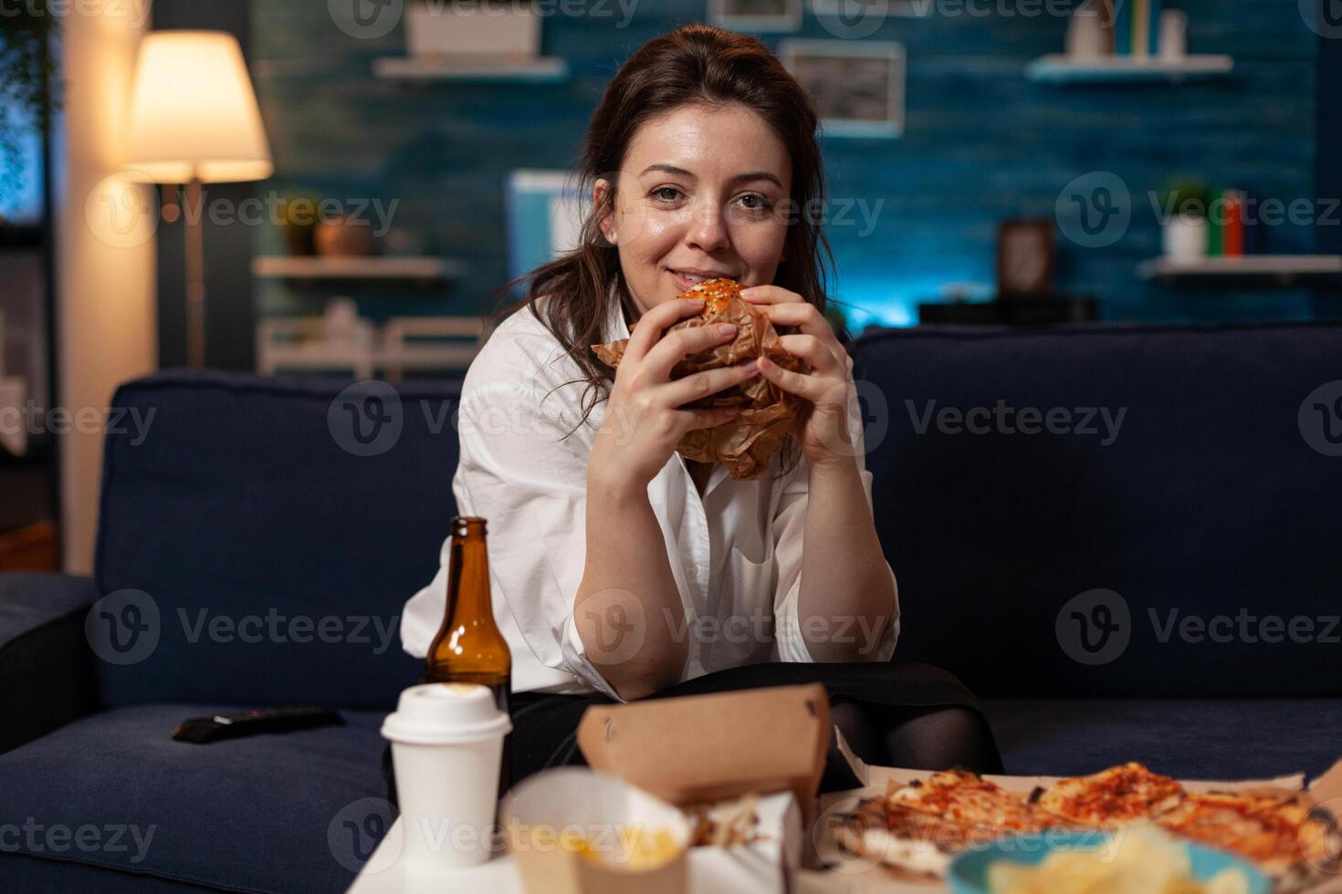 Smiling woman holding tasty burger in hands looking at television comedy sitcom standing on couch in front of table with takeaway fast food menu. Happy person watching tv show having takeout dinner. photo